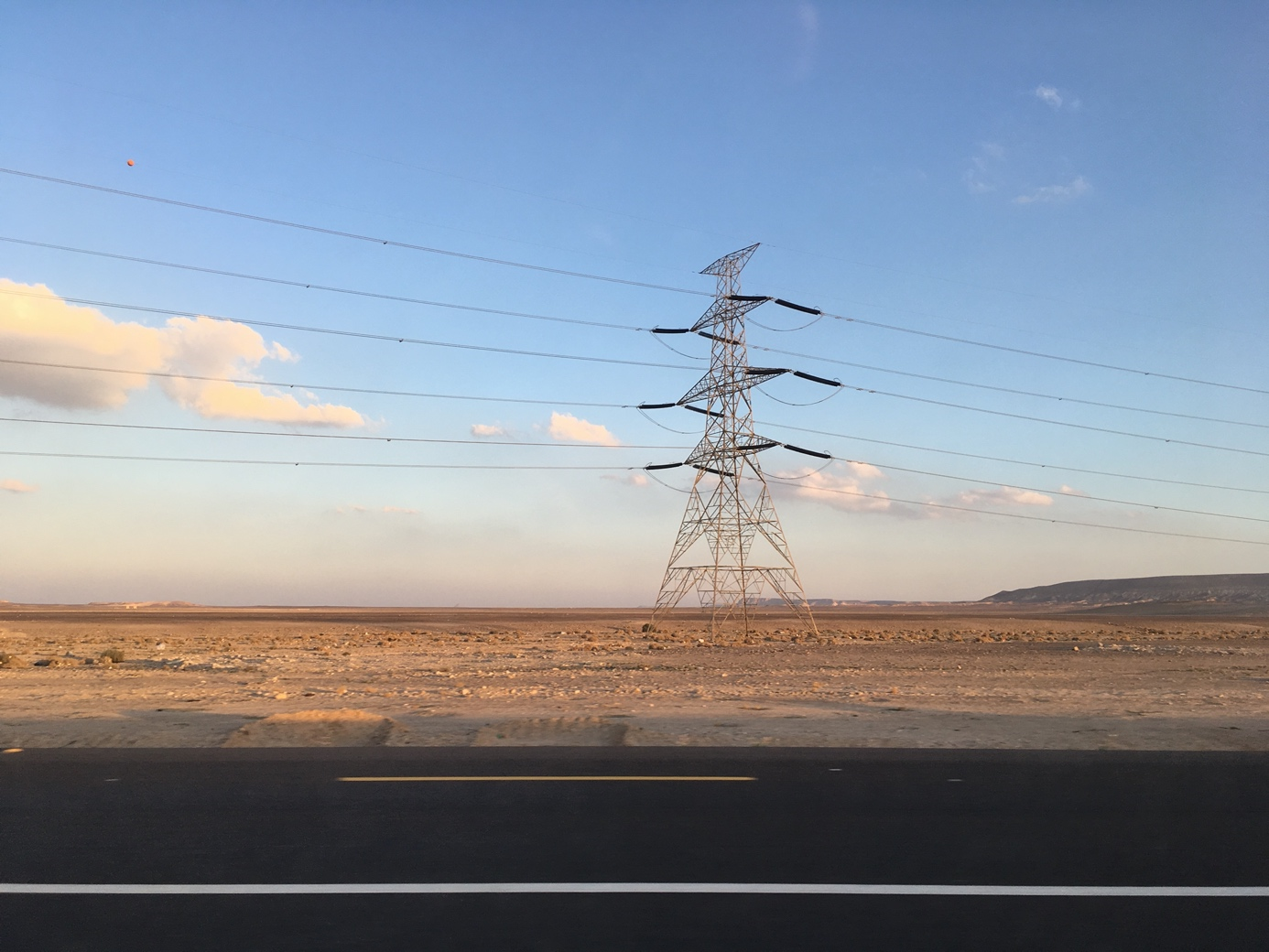 Das Foto zeigt einen Strommast und Leitungen vor blauem Himmel in einer Wüstenlandschaft. Im Vordergrund ist eine Straße zu sehen.