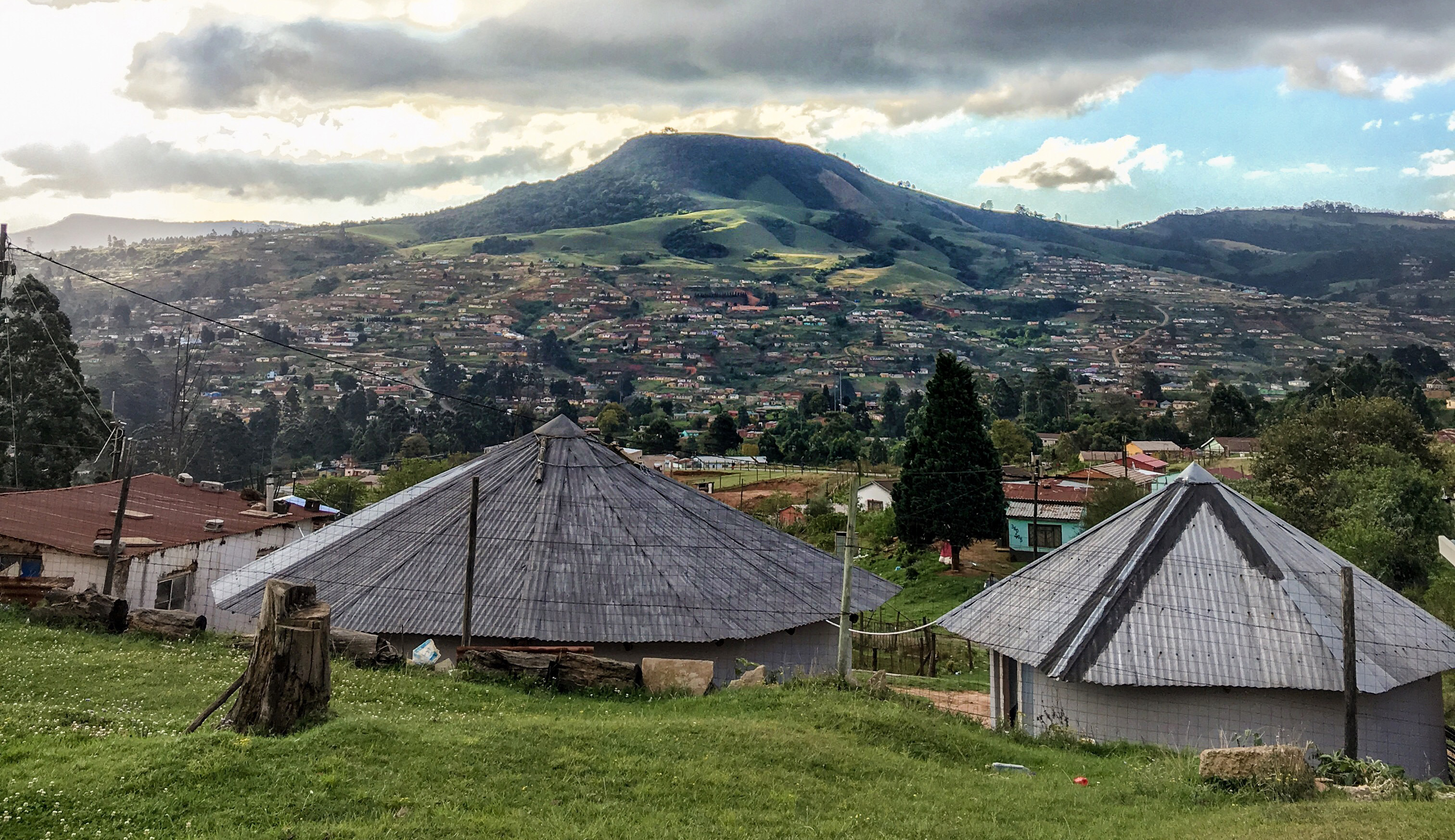 Das Bild zeigt eine hügelige Landschaft. Eine Siedlung bedeckt teilweise die Hänge. Die Hügelkuppen sind teilweise mit Wald bedeckt. Im Vordergrund befinden sich zwei traditionelle Zulu-Hütten (Rundhütten mit Spitzdächern) und eine Wohnhütte..