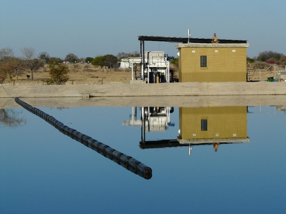 Im Vordergrund Wasseroberfläche des ersten Abwasserteiches mit installierter schwimmender Leitwand. Im Hintergrund Vorbehandlung mit Mikrosieb und Schaltwarte.