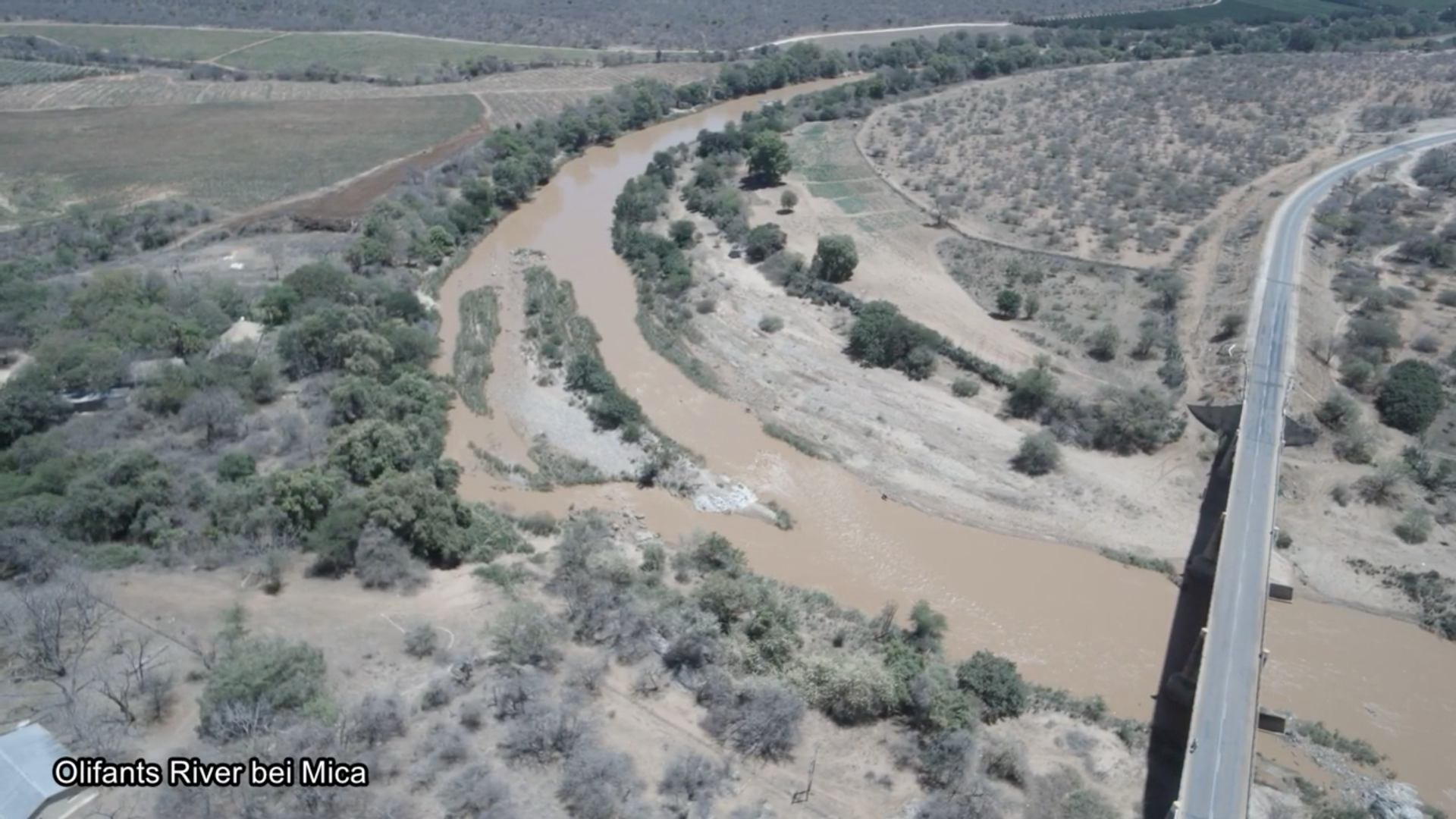 Es ist eine Luftaufnahme eines Flusses und seines Umlandes zu sehen. Über den Fluss führt eine betonierte Brücke. Das Flusswasser ist braun, die Erde des Umlandes besitzt ebenfalls eine braune Färbung und wird von einigen grünen Bäumen und Büschen durchzogen. Unten links im Bild steht: Olifants River bei Mica.