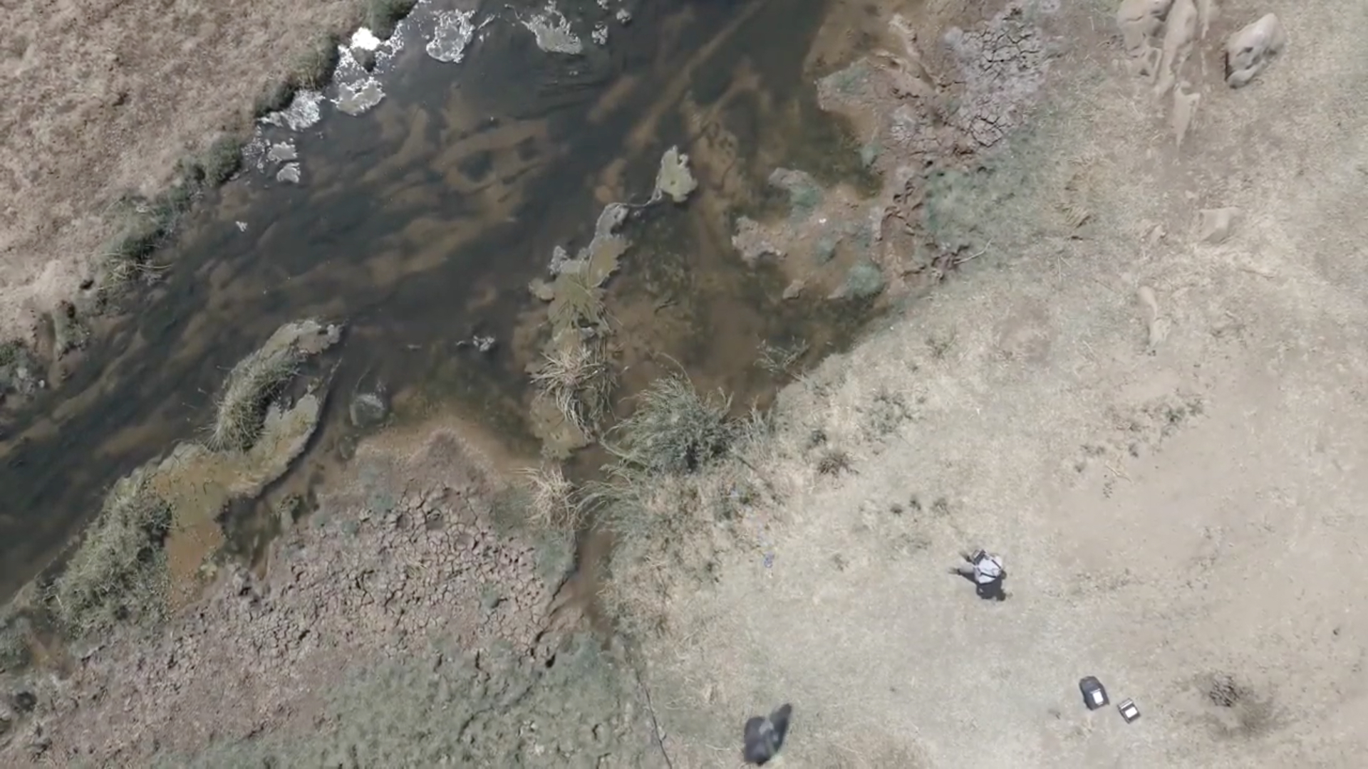 Zu sehen ist eine Luftaufnahme eines Flusses aus der Drohnenperspektive. An der Drohne hängt eine Vorrichtung, die eine Wasserprobe aus dem Fluss entnehmen soll.