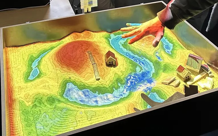 m Sand sind Landschaftsformen wie Hügel und Täler modelliert, deren Höhenunterschiede durch eine farbige Projektion hervorgehoben werden. Das projizierte Geländemodell zeigt einen relativen Farbverlauf von Blau (unter dem Meeresspiegelniveau) über Grün, Gelb und Orange bis hin zu Rot und Braun für höher gelegene Bereiche. Die höchsten Darstellungsbereiche werden Weiß eingefärbt. Über dem Sand schwebt eine Hand, mit der Regen simuliert werden kann. Auf der Sandfläche ist zudem eine Wasseroberfläche sichtbar, die den simulierten Wasserfluss über das Gelände zeigt.